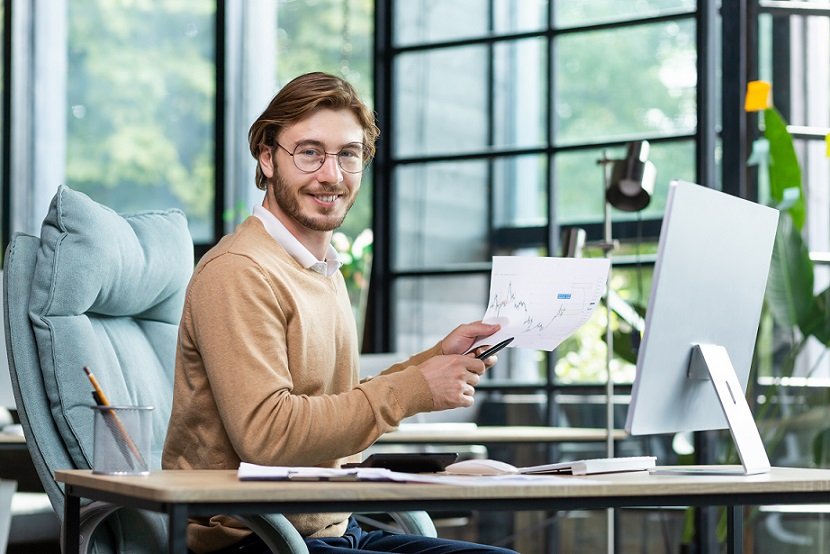 Smiling young boss sits at a desk in front of a computer, makes a video call presentation online, shows graphs, looks at the camera and smiles.